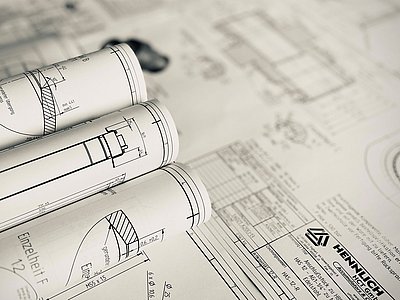 Eine technische Zeichnung liegt ausgebreitet im Hintergrund. Darauf liegen drei zusammengerollte Zeichnungen